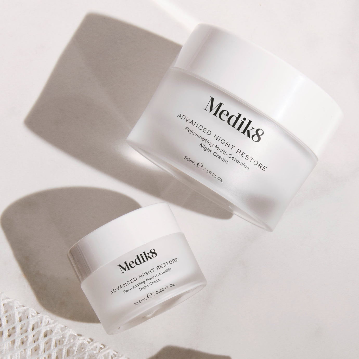 Two white Medik8 Advanced Night Restore cream jars, one 50ml full size and one 12.5ml travel size, set against a white textured background with sharp diagonal shadows and a piece of white netting. Text on the jars reads 