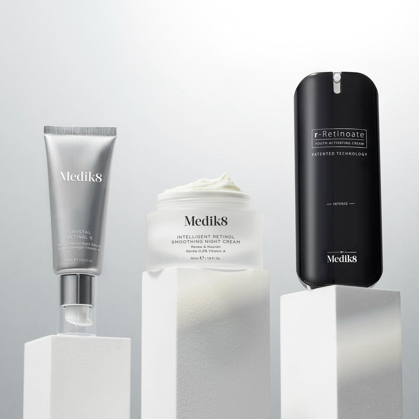 Three Medik8 vitamin A products displayed on white plinths against a light grey background. From left: a silver Crystal Retinal 6 pump tube, an open white Intelligent Retinol Smoothing Night Cream jar revealing its texture, and a black r-Retinoate Intense pump bottle. Text highlights key claims for each.