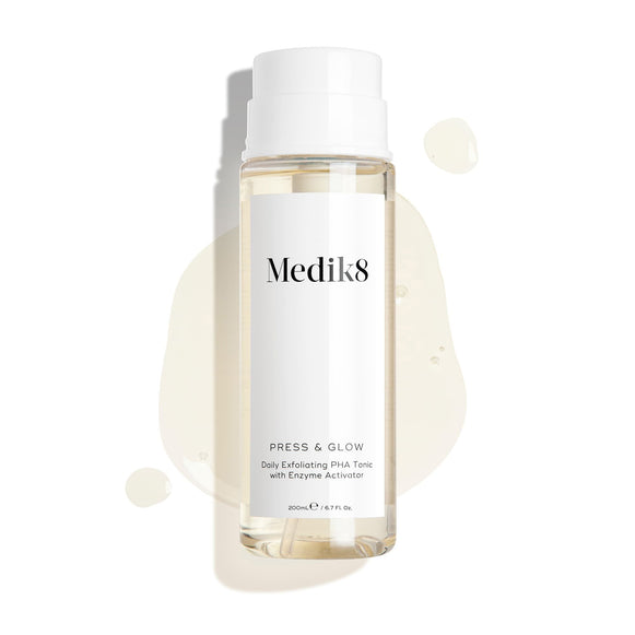 Medik8 Press & Glow tonic bottle stands against a clean white background with translucent liquid droplets and a puddle, highlighting its watery texture. Text identifies the product as a 200mL "Daily Exfoliating PHA Tonic with Enzyme Activator."-7