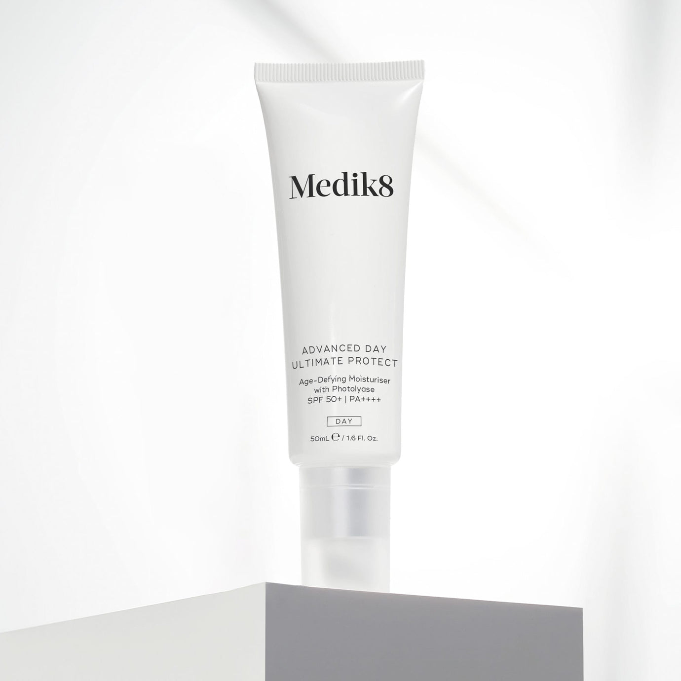 A white 50mL tube of Medik8 Advanced Day Ultimate Protect cream standing upright on a light grey geometric block. Set against a soft, draped white fabric background, the text on the tube highlights it as an 