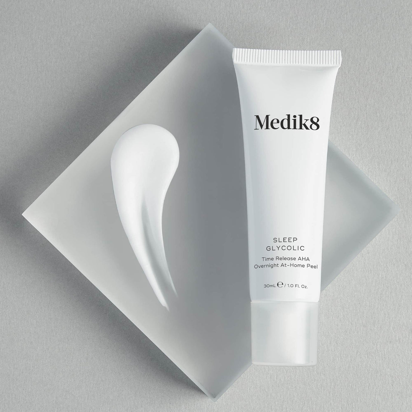 A white Medik8 Sleep Glycolic tube resting on a frosted, translucent square block against a textured light grey background. A smooth swoosh of white cream sits beside the tube. Text details it as a 