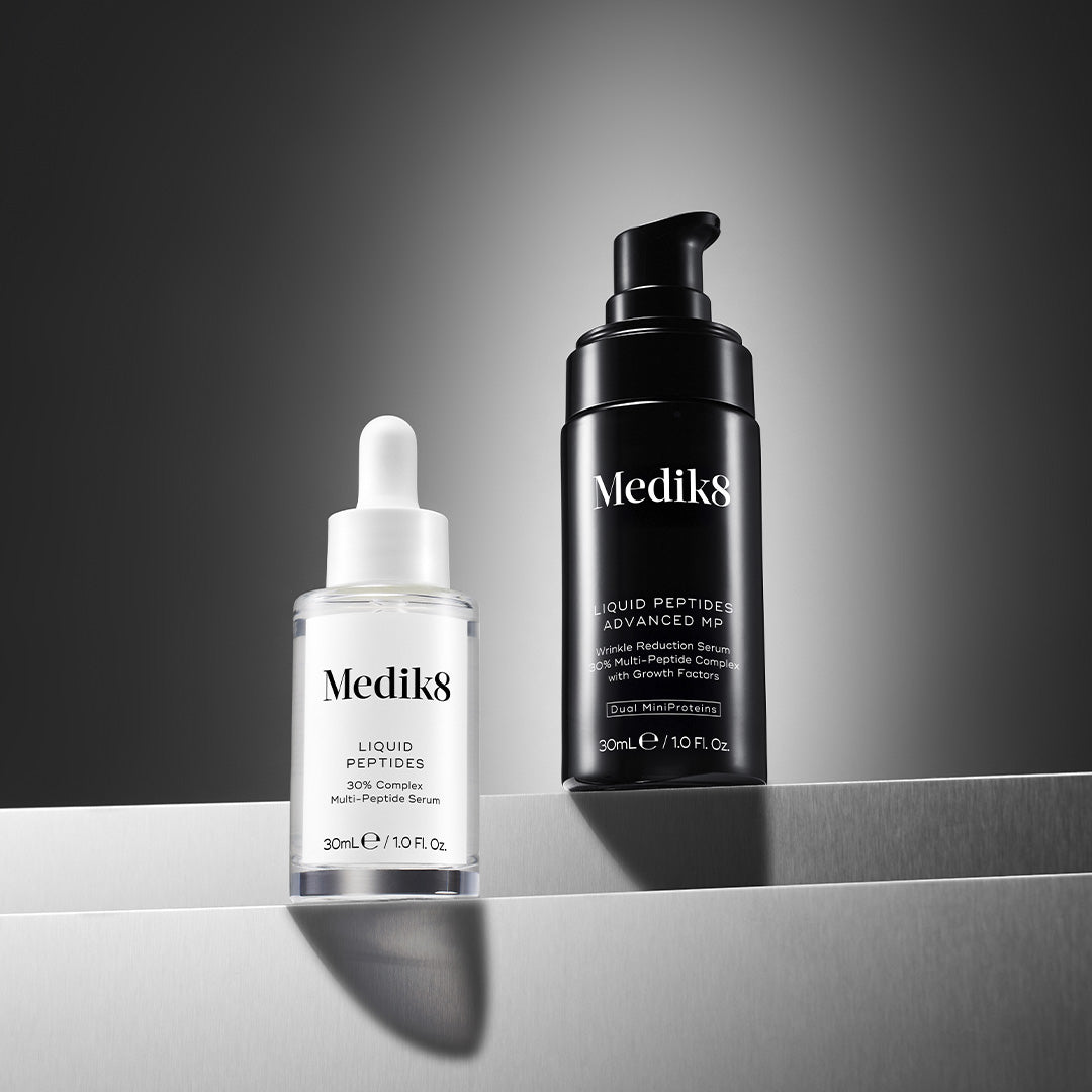 Two Medik8 serums displayed on silver metallic steps against a dark grey gradient background. On the left is a clear Liquid Peptides dropper bottle (30% Complex Multi-Peptide Serum). On the right is a tall black Liquid Peptides Advanced MP pump bottle featuring text: 