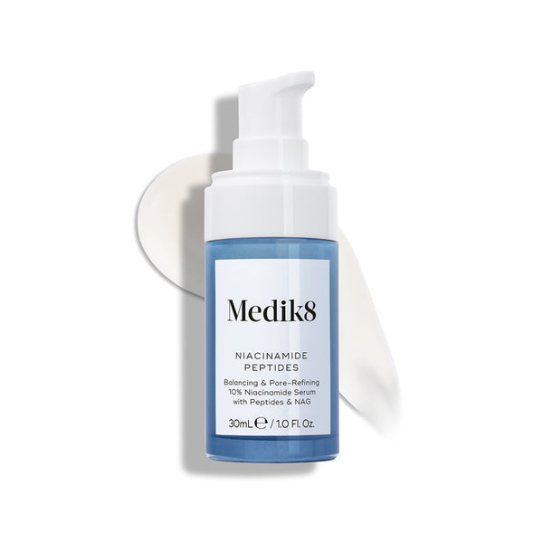 Medik8 Niacinamide Peptides serum bottle with a white pump top, resting on a white surface with a smear of white serum behind it.