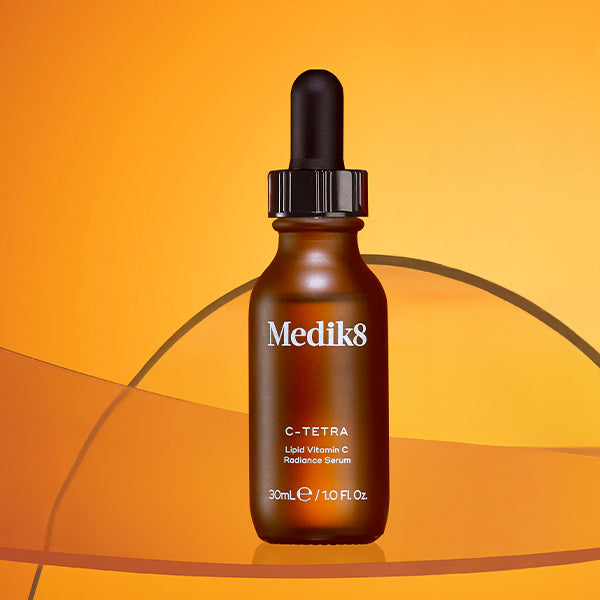 A Medik8 C-Tetra Lipid Vitamin C Radiance Serum dropper bottle displayed against a warm orange gradient background. A translucent, sweeping curved orange graphic element crosses behind and beneath the glass bottle.