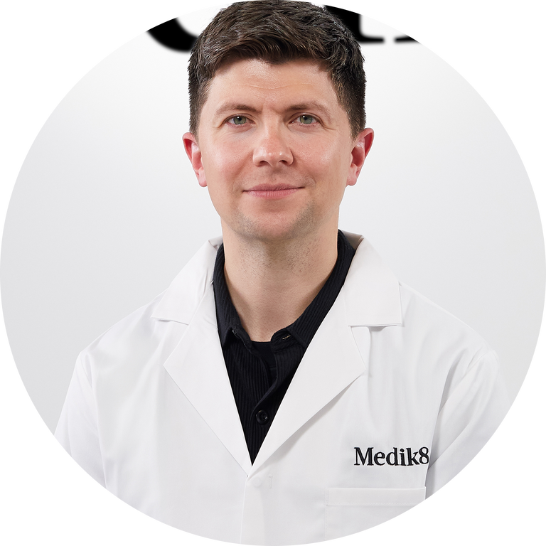 A circular headshot portrait of a man with light brown hair and green eyes, smiling neutrally. He is wearing a white lab coat with 'Medik8' embroidered on the pocket and a black button-up shirt. The background is white.