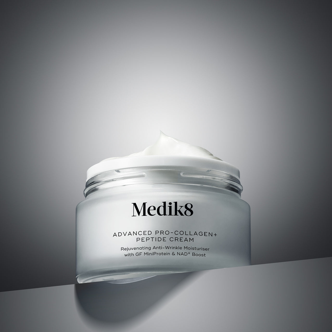 A professional photograph of an open jar of Advanced Pro-Collagen Peptide Cream, revealing its smooth white texture, resting on a grey geometric surface against a grey gradient background. Below the image, clear text identifies this product category as ‘Moisturisers’.