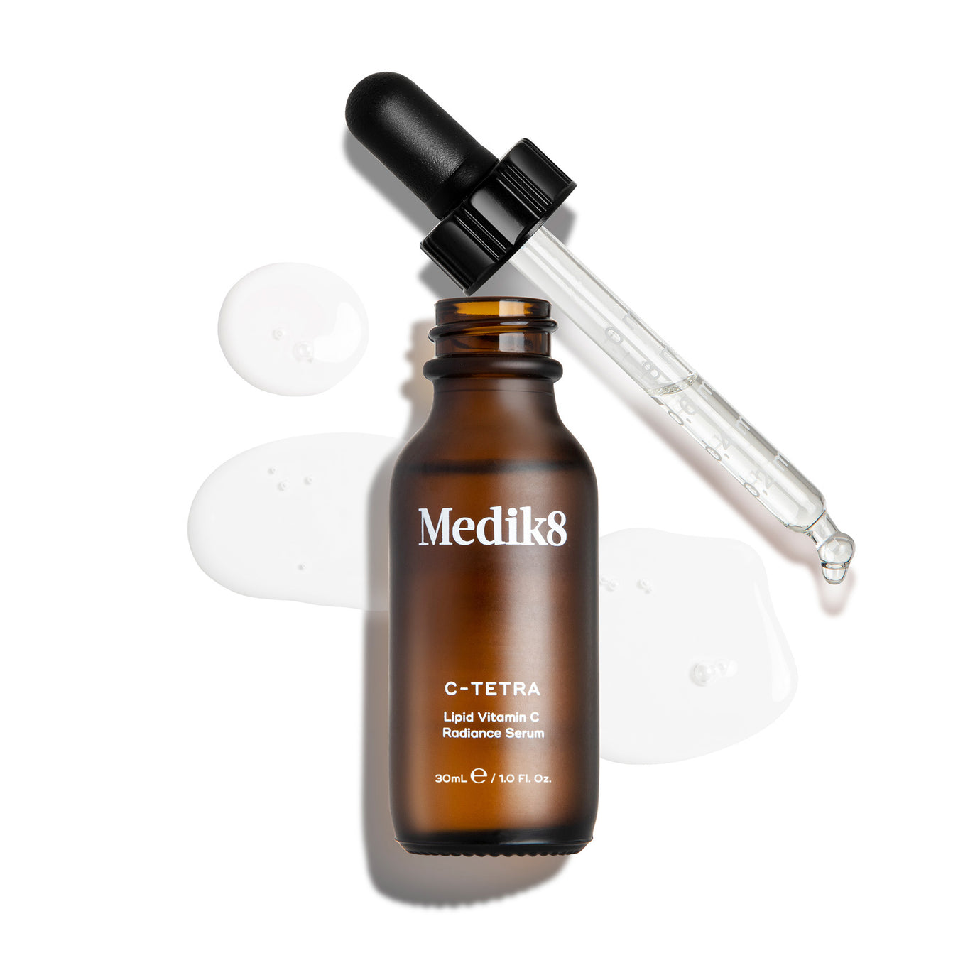 A bottle of Medik8 C-Tetra Lipid Vitamin C Radiance Serum open with its dropper resting diagonally across the top. Set against a clean white background, the bottle is surrounded by three clear puddles of serum, showcasing its liquid texture.