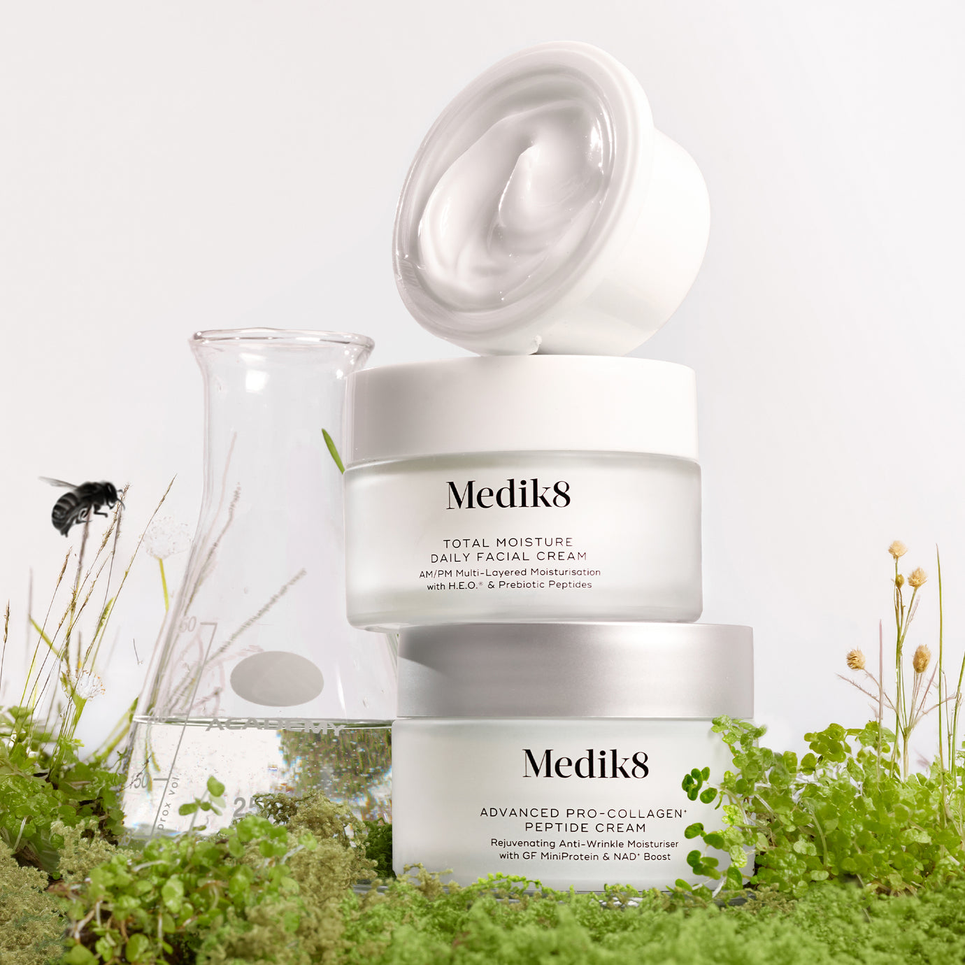 A stack of Medik8 moisturizers and a laboratory beaker placed amongst lush green moss and wildflowers, symbolizing the harmony between clinical science and environmental responsibility.