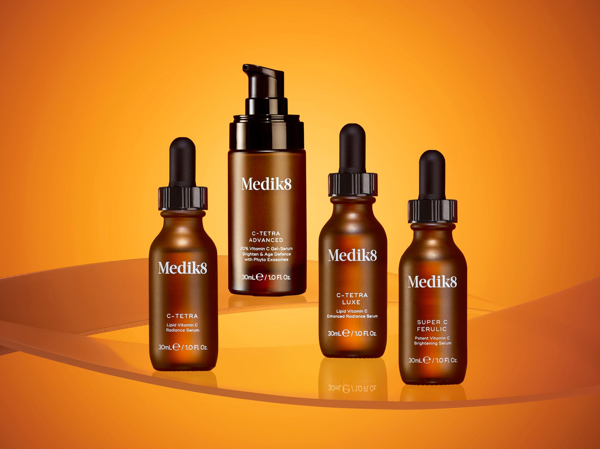 A family of four Medik8 Vitamin C serums displayed on a reflective orange surface against a vibrant orange background. The lineup includes amber glass bottles of C-Tetra, C-Tetra Luxe, and Super C Ferulic with dropper caps, alongside a larger pump bottle of C-Tetra Advanced. The warm, sun-drenched lighting emphasizes the products' focus on skin radiance and antioxidant protection.