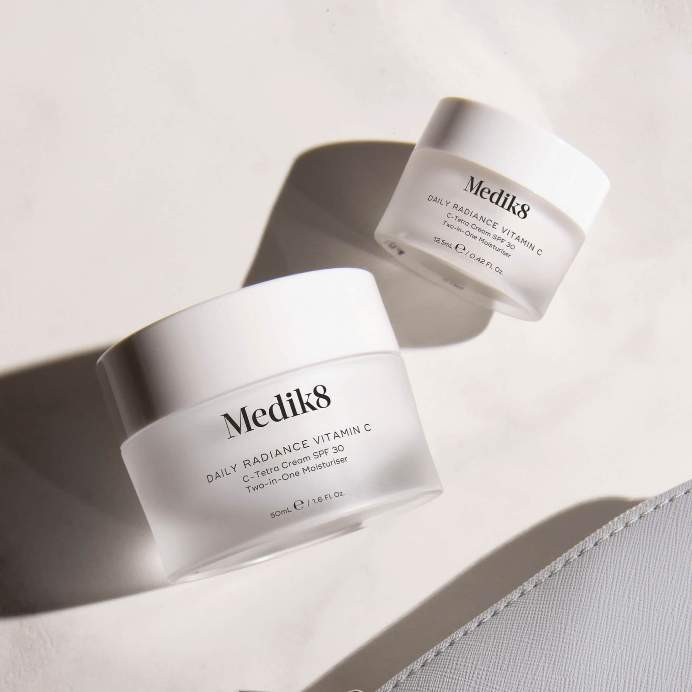Two jars of Medik8 Daily Radiance Vitamin C cream displayed on a white surface with shadows. The larger jar (50ml) is in the foreground, and a travel-size jar (12.5ml) is in the background. Text identifies it as 