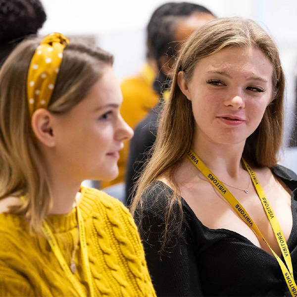 Two young women are seated in a lecture hall or event space, engaged in conversation. One woman wears a yellow top and a matching yellow polka-dot headband, while the other wears a black top and a yellow lanyard. 