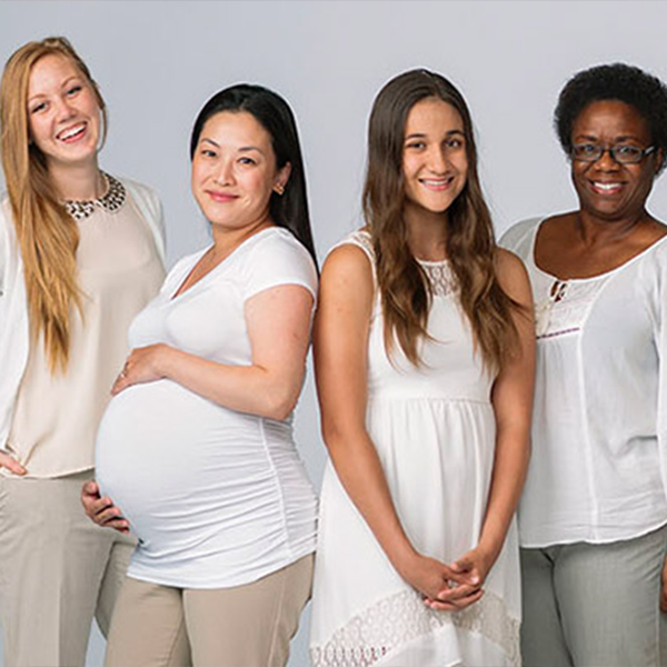 Four smiling women standing side-by-side against a plain light grey background. From left to right: a laughing woman with long blonde hair in a cream top; a pregnant woman with long dark hair holding her bump in a fitted white t-shirt; a woman with long brown hair in a white sleeveless dress; and a woman with short dark curly hair and glasses in a white blouse. All are dressed in light-coloured clothing.